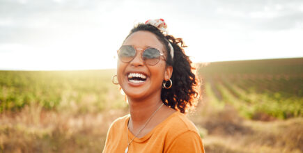 A woman stands in a field. She is smiling and wearing sunglasses.
