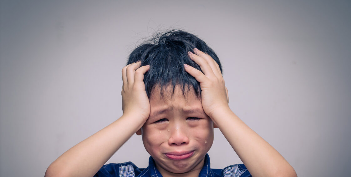 A young Asian boy holds is head and looks directly ahead while crying. There are visible tears on his cheeks.