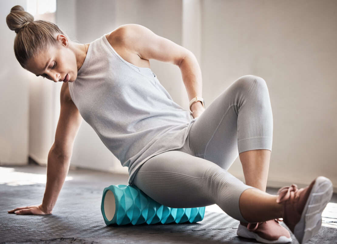 A white woman wearing a gray tanktop and workout leggings is foam rolling her right glute on a blue foam roller