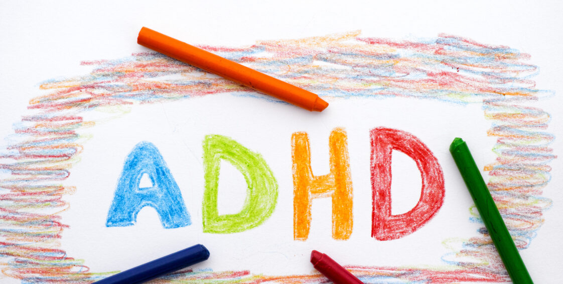 The words "ADHD" are written in different colors using crayon. There are orange, blue, red, and green crayons laying on top of the paper.