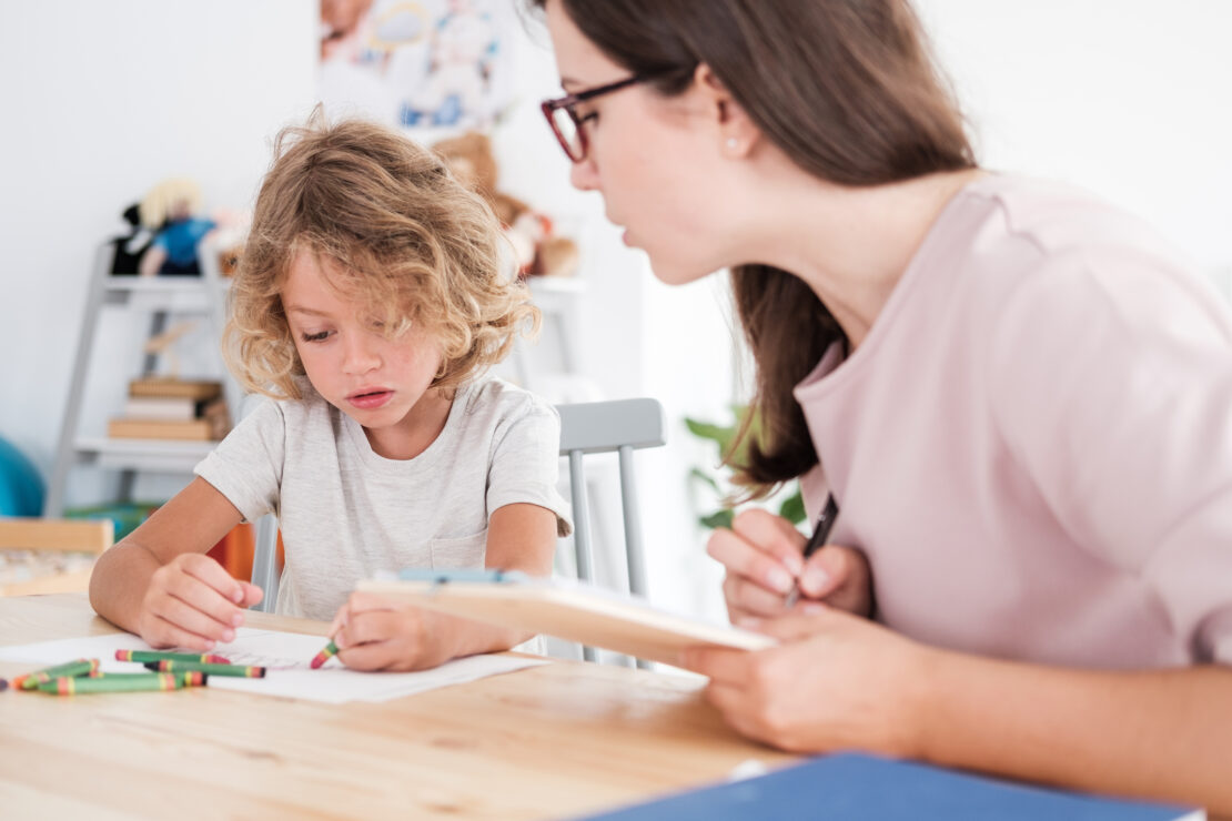 A young boy draws with crayon while a female therapist watches. The therapist is holding a pen and clipboard to take notes.