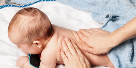 A small, five-month-old baby is laying on a soft blanket. An unknown person has their hands placed on the baby's back, performing a chiropractic adjustment.