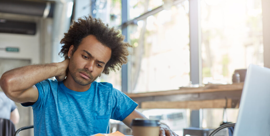 A young man sits at a desk. He is looking at his laptop and has his hand placed on the back of his neck. He is wearing an expression of discomfort.